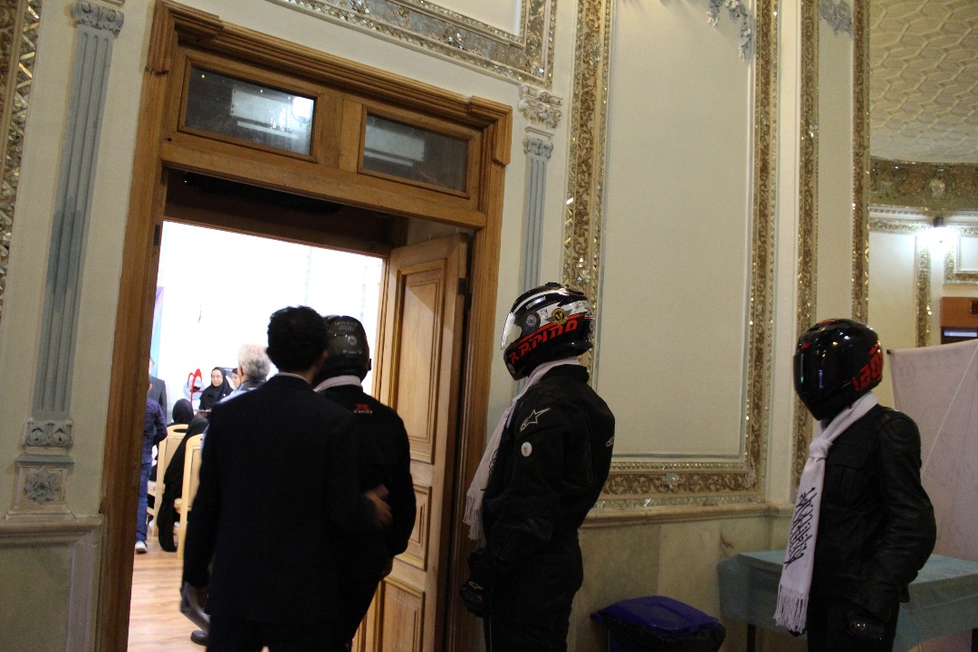 3 موتورسوار با لباس و کلاه موتورسواری که دور گردنشان شال سفید پیچیده شده در حال وارد شدن از چارچوب چوبیه در به سالن برگزاری مراسم اهدا عضو