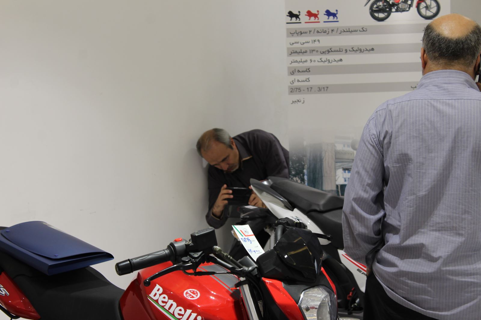 تصویری از یک موتورسیکلت بنللی قرمز رنگ در نمایشگاه موتورسیکلت در حالی که مردی در کنار آن خم شده تا از آن عکس بگیرد.