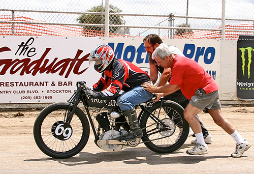 یک موتورسوار مرد با لباس و کلاه موتوروساری به رنگ قرمز و مشکی سوار بر موتورسیکلت سیاه رنگ که دو مرد یکی میانسال با تیشرت قرمز و شلوارک آبی و مرد جوانی در کنارش در حال هل دادن موتورسیکلت هستند